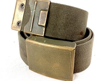 May include: An olive green canvas belt featuring a rectangular brass buckle with a textured stippled surface. The belt includes a secondary brass metal attachment piece with two circular eyelets, showing a weathered finish on the metal components.