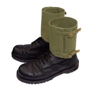 May include: A pair of black leather boots with olive green canvas gaiters. The boots have black laces and a rounded toe. The gaiters are secured with two brass-coloured buckles and straps.