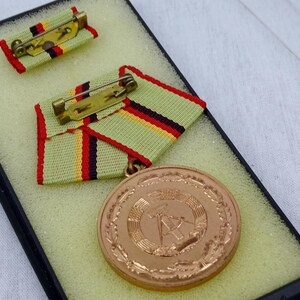 May include: A bronze medal with a wreath design and a hammer and compass in the center. The medal is displayed in a black box with a green and yellow ribbon.
