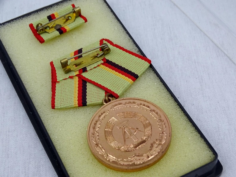 May include: A bronze medal with a wreath design and a hammer and compass in the center. The medal is displayed in a black box with a green and yellow ribbon.