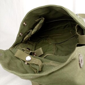 May include: A green canvas bag with a flap closure and multiple straps and buckles. The bag has a large main compartment and a smaller pocket on the inside.