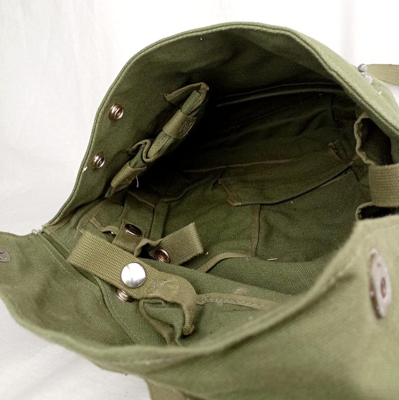 May include: A green canvas bag with a flap closure and multiple straps and buckles. The bag has a large main compartment and a smaller pocket on the inside.