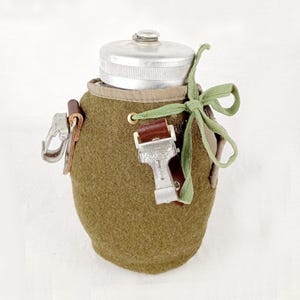 Puede incluir: Una cantimplora verde oliva con tapa plateada y lazo verde. La cantimplora tiene una correa de cuero marrón y un cierre metálico. La cantimplora está cubierta con un material similar al fieltro.