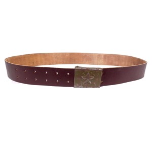 May include: A dark red leather belt with a metal buckle featuring a five-pointed star. The belt has a series of evenly spaced holes for adjusting the fit.