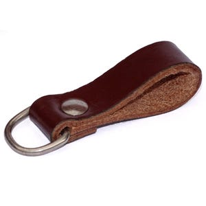 Op de afbeelding: Een bruine leren sleutelhanger met een zilveren metalen ring. De sleutelhanger heeft een enkele steek en een metalen knoop.