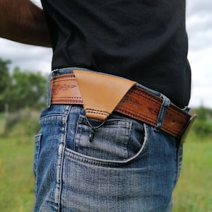 Puede incluir: Un cinturón de cuero marrón con un intrincado diseño floral. El cinturón se lleva alrededor de la cintura de una persona que lleva pantalones vaqueros azules. Una funda de cuero marrón está unida al cinturón.