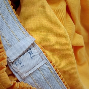 Pu&ograve; includere: Primo piano di un paio di pantaloncini gialli con una fascia elastica bianca. La fascia ha un'etichetta con la scritta "SPORT USA". I pantaloncini sono realizzati in un tessuto strutturato e hanno un design a pieghe.