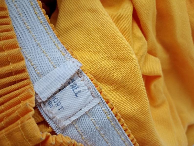 Pu&ograve; includere: Primo piano di un paio di pantaloncini gialli con una fascia elastica bianca. La fascia ha un'etichetta con la scritta "SPORT USA". I pantaloncini sono realizzati in un tessuto strutturato e hanno un design a pieghe.