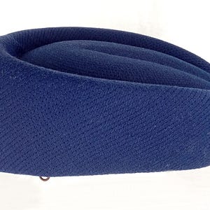 Pu&ograve; includere: Un cappello blu navy con un design curvo e arrotolato. Il cappello &egrave; realizzato in un tessuto strutturato e ha un piccolo anello di metallo sul fondo. La forma del cappello ricorda un tradizionale berretto militare o di servizio.