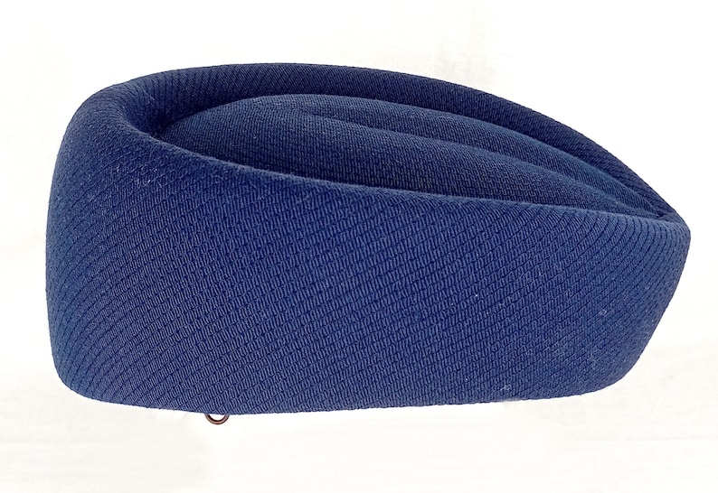 Pu&ograve; includere: Un cappello blu navy con un design curvo e arrotolato. Il cappello &egrave; realizzato in un tessuto strutturato e ha un piccolo anello di metallo sul fondo. La forma del cappello ricorda un tradizionale berretto militare o di servizio.