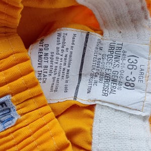 Pu&ograve; includere: Primo piano di pantaloncini sportivi giallo brillante con un'etichetta bianca. L'etichetta recita "TRUNKS, GENERAL PURPOSE: EXERCISER" e include la taglia (36-38). I pantaloncini hanno una piccola etichetta blu con "FOOD".