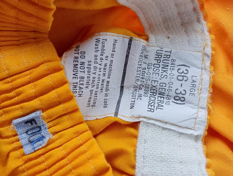 Pu&ograve; includere: Primo piano di pantaloncini sportivi giallo brillante con un'etichetta bianca. L'etichetta recita "TRUNKS, GENERAL PURPOSE: EXERCISER" e include la taglia (36-38). I pantaloncini hanno una piccola etichetta blu con "FOOD".
