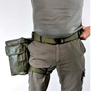 Puede incluir: Un cinturón utilitario verde oliva con una bolsa grande y un bolsillo más pequeño. El cinturón tiene una hebilla negra y una correa verde a juego que se asegura alrededor del muslo. La persona lleva pantalones verde oliva y una camisa a juego.