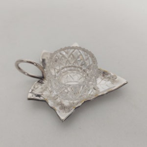 Può includere: Un saliera in vetro trasparente con un design in vetro tagliato si trova su un vassoio d'argento con un design a foglia. Il vassoio ha un manico.