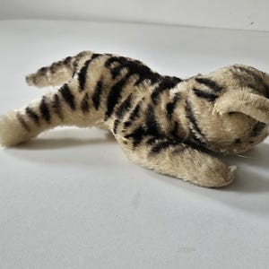 Op de afbeelding: Een liggende pluche kat. De kat heeft een beige lichaam met zwarte strepen, en de poten en oren zijn lichtbeige. Het speelgoed is gemaakt van een zacht, pluizig materiaal.
