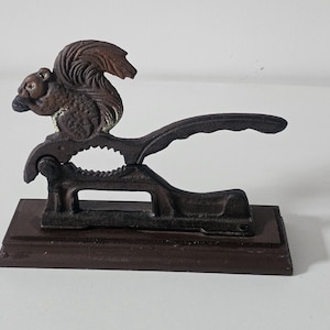 Könnte beinhalten: Ein Nussknacker aus Gusseisen mit einem Eichhörnchen-Design. Das Eichhörnchen sitzt auf dem Hebel des Nussknackers, der an einer Basis befestigt ist. Der Nussknacker ist braun und hat ein rustikales Aussehen, passend für die Kücheneinrichtung.