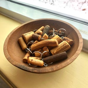 Puede incluir: Un cuenco de madera lleno de varios llaveros de madera. Los llaveros están hechos de pequeños trozos cilíndricos de madera con anillos metálicos. El cuenco es de madera marrón claro y está sobre una superficie amarilla.