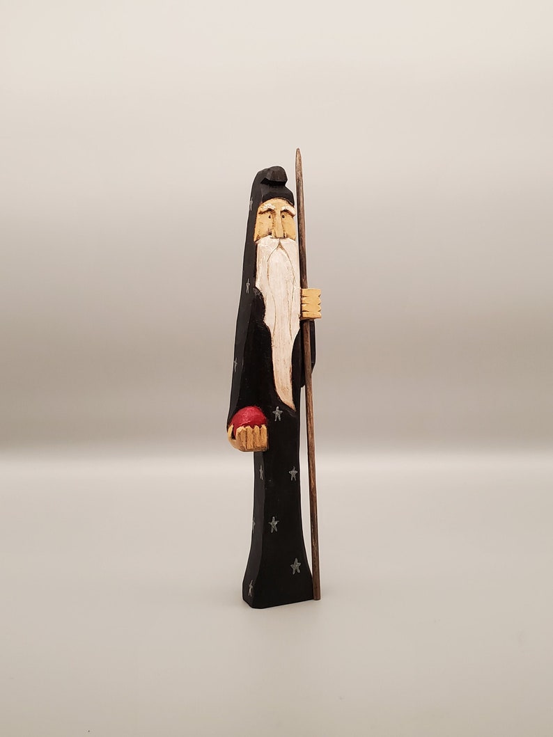 Puede incluir: Una figurita de madera de un mago con una larga barba blanca, vistiendo una t&uacute;nica negra con estrellas plateadas y sosteniendo un orbe rojo en una mano y un bast&oacute;n en la otra.
