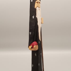 Puede incluir: Una figurita de madera tallada de un mago con una larga barba blanca, vistiendo una t&uacute;nica negra con estrellas plateadas, sosteniendo un orbe rojo en su mano izquierda y un bast&oacute;n de madera en su mano derecha.