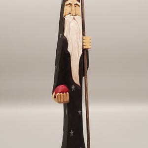 Puede incluir: Una figurita de madera tallada de un mago con una larga barba blanca, vistiendo una t&uacute;nica negra con estrellas plateadas, sosteniendo un orbe rojo y un bast&oacute;n de madera.