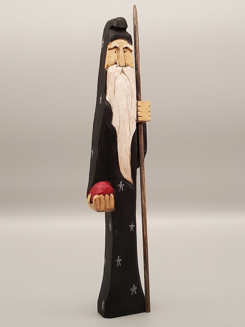 Puede incluir: Una figurita de madera tallada de un mago con una larga barba blanca, vistiendo una t&uacute;nica negra con estrellas plateadas, sosteniendo un orbe rojo y un bast&oacute;n de madera.