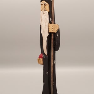 Puede incluir: Una figurita de madera tallada de un mago con una larga barba blanca, vistiendo una t&uacute;nica negra con estrellas plateadas, sosteniendo un bast&oacute;n y una cesta roja.