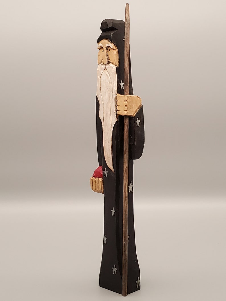 Puede incluir: Una figurita de madera tallada de un mago con una larga barba blanca, vistiendo una t&uacute;nica negra con estrellas plateadas, sosteniendo un bast&oacute;n y una cesta roja.