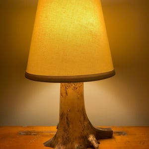 May include: A rustic table lamp with a natural tree trunk base and a beige lampshade. The lampshade has a dark brown trim. The lamp is sitting on a wooden table. The lamp is lit, casting a warm glow.