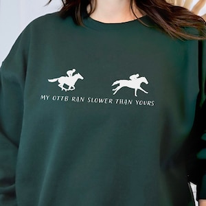 May include: Dark green sweatshirt with white graphic of two racehorses and riders. Text reads "MY OTTB RAN SLOWER THAN YOURS". The sweatshirt has a crew neck and long sleeves.
