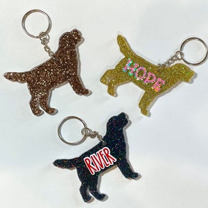 May include: Three glittery dog-shaped keychains. One is brown, one is gold with the word "HOPE", and one is black with the word "RIVER". Each keychain has a silver keyring attached with a chain. The keychains are on a white background.