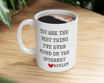 Op de afbeelding: Witte keramische koffiemok met een zwart interieur, gevuld met koffie. De mok heeft de tekst "YOU ARE THE BEST THING I'VE EVER FOUND ON THE INTERNET" met een rood hart en de naam "Ashley".