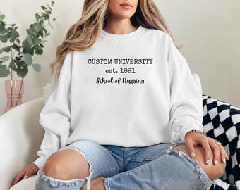 Pode incluir: Camisola branca com o texto "CUSTOM UNIVERSITY est. 1891 School of Nursing" em preto. A camisola é usada por uma pessoa que usa jeans azuis. A pessoa está sentada numa cadeira branca.