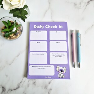 Peut inclure: Un bloc-notes violet clair "Daily Check In" avec des sections pour l'hygiène, les tâches, les repas et autres. Comprend un dessin de koala et le texte "Cerulean". Deux stylos, un rose et un bleu, sont à côté du bloc-notes.