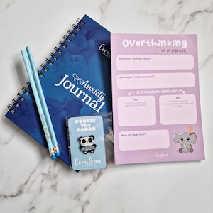 Peut inclure: Un ensemble d'articles de soins personnels, comprenant un "Anxiety Journal" bleu à spirales, deux crayons bleu clair, une épingle "Pookie the Panda" et une feuille de travail rose "Overthinking in progress". Le journal et la feuille de travail ont le logo "Cerulean Journals".