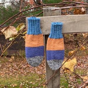 May include: Two knitted mittens in blue, orange, purple, and grey. They are hanging on a wooden fence post outdoors, with autumn leaves in the background. These mittens are a warm accessory for cold weather, perfect for winter.