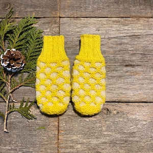 May include: A pair of yellow knitted mittens with a white honeycomb pattern. The mittens are displayed on a rustic wooden surface, with a sprig of evergreen and a pinecone in the background. The mittens are a winter accessory.