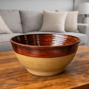 May include: A large, round ceramic bowl featuring a two-tone design. The upper portion has a glossy, deep reddish-brown glaze with visible horizontal ridges, while the lower portion has a matte, light tan finish with a textured, earthy appearance.