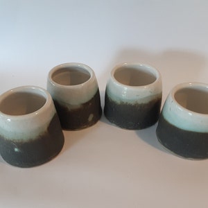 May include: Four small ceramic cups with a white glaze and a brown glaze band around the bottom. The cups are arranged in a row on a white surface.
