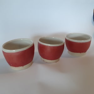May include: Three small ceramic bowls with a white rim and a red glaze. The bowls are arranged in a row on a white surface.