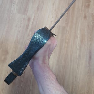 May include: A dark, antique-style dagger with a textured, hammered metal handle and a long, slender blade. The handle is a dark, matte color, and the blade is silver. The dagger is held in a hand.