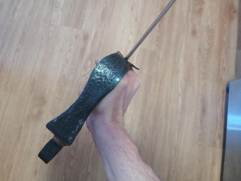 May include: A dark, antique-style dagger with a textured, hammered metal handle and a long, slender blade. The handle is a dark, matte color, and the blade is silver. The dagger is held in a hand.