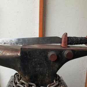 May include: A black metal anvil with a rusty surface and a chain wrapped around the base. A handmade knife with a black leather wrapped handle and a wooden pommel is resting on the anvil.