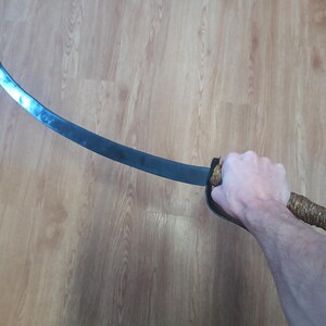 May include: A curved, dark metal sword with a wrapped handle and a decorative pommel. The blade has a slight curve and a dull finish. The handle is wrapped in a textured, light brown material.