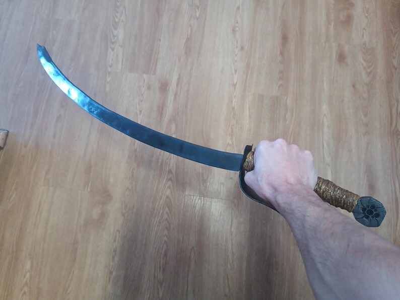 May include: A curved, dark metal sword with a wrapped handle and a decorative pommel. The blade has a slight curve and a dull finish. The handle is wrapped in a textured, light brown material.