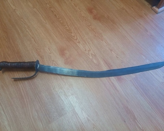 May include: A curved, silver-colored sword with a brown leather-wrapped handle. The sword is lying on a wooden surface.