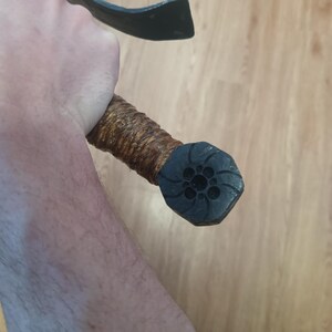 May include: A close-up of a black and brown axe. The axe head is black, and the handle is wrapped in brown material. The end of the handle has a black, octagonal cap with a floral design.