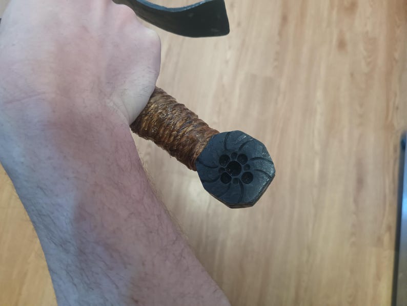 May include: A close-up of a black and brown axe. The axe head is black, and the handle is wrapped in brown material. The end of the handle has a black, octagonal cap with a floral design.