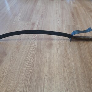 May include: A curved, black metal sword with a textured, brown handle and a guard. The blade is approximately 30 inches long. The sword is lying on a wooden surface.