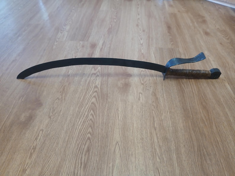 May include: A curved, black metal sword with a textured, brown handle and a guard. The blade is approximately 30 inches long. The sword is lying on a wooden surface.