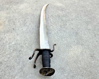 May include: A curved metal sword featuring a long, tapered blade with a sharp point. The handle is wrapped in black cord and includes a dark metal crossguard with curved quillons and a circular pommel at the base of the grip.